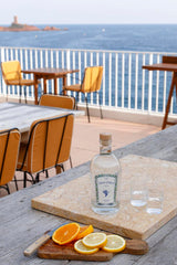 Flasche Ziga Zaga Vodka mit zwei Gläsern und Orangenscheiben auf Terrasse mit Meerblick und Tischen im Hintergrund.