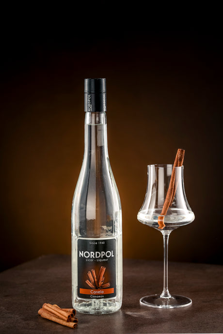 Flasche Nordpol Zimtlikör (Canela) neben Glas mit Zimtstange als Garnitur, vor dunkelbraunem Hintergrund, zusätzliche Zimtstangen vorne.