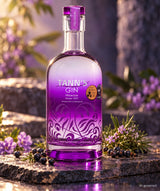 Tann's Premium Gin in violetter Flasche auf einer Steinplatte mit Wacholderbeeren, Kräutern und violetten Blüten.